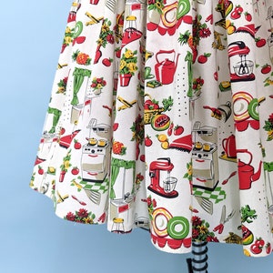 May include: A white cotton fabric with a red, green and yellow kitchen-themed print. The print features images of kitchen appliances, food and flowers.