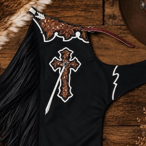 May include: A black leather chap featuring long fringe along the side, adorned with a brown floral embossed leather yoke and a matching cross applique. The design includes white piping accents and a thin adjustable strap with a metal buckle.