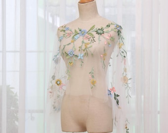 May include: A sheer, off-white mesh capelet featuring delicate, multi-colored floral embroidery. The design includes small blue, yellow, pink, and green flowers with trailing vines and leaves scattered across the translucent fabric, draped over a mannequin.
