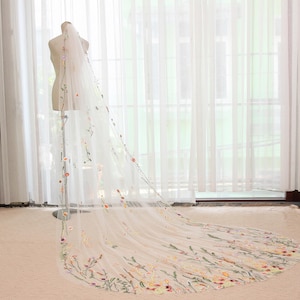 May include: A long, sheer white bridal veil with delicate floral embroidery in shades of orange, yellow, and green. The veil is displayed on a mannequin, showcasing its length and intricate design. The floral pattern is concentrated at the edges.