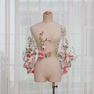May include: A sheer, white top with long sleeves, adorned with embroidered pink and peach flowers and green leaves. The floral design is concentrated on the front and sleeves of the garment, creating a delicate and feminine aesthetic.