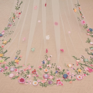 Puede incluir: Un velo transparente de color marfil adornado con flores bordadas de colores. El diseño floral incluye flores rosas, azules, amarillas y blancas con hojas verdes, creando una estética delicada y romántica. Adecuado para bodas u ocasiones especiales.