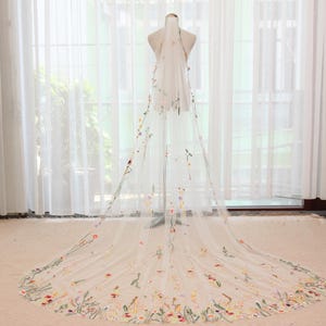 May include: A long, sheer white wedding veil with delicate floral embroidery in shades of pink, yellow, and green. The veil is displayed on a mannequin, showcasing its intricate design and flowing fabric. The floral details are concentrated along the edges.