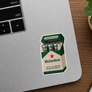 Peut inclure: Un autocollant repr&eacute;sentant une bo&icirc;te de cigarettes avec la marque Heineken, &eacute;tiquet&eacute;e "FRIDGE CIGS". La bo&icirc;te est verte et blanche, avec une &eacute;toile rouge et le texte "JUST A LITTLE SOMETHING TO TAKE THE EDGE OFF". L'autocollant est sur un ordinateur portable.