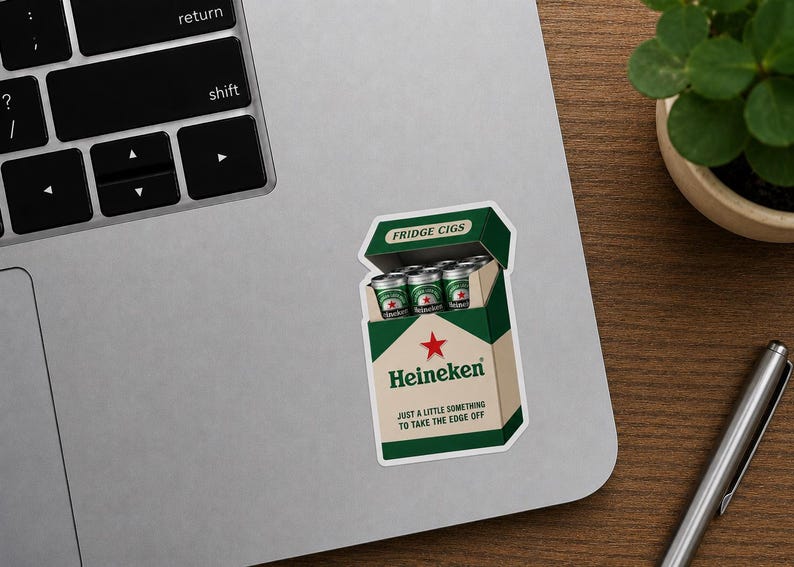 Peut inclure: Un autocollant repr&eacute;sentant une bo&icirc;te de cigarettes avec la marque Heineken, &eacute;tiquet&eacute;e "FRIDGE CIGS". La bo&icirc;te est verte et blanche, avec une &eacute;toile rouge et le texte "JUST A LITTLE SOMETHING TO TAKE THE EDGE OFF". L'autocollant est sur un ordinateur portable.