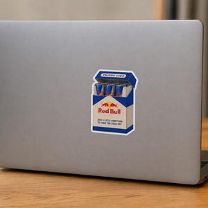 May include: A rectangular sticker on a laptop lid featuring a graphic of a blue and white cigarette pack labelled FRIDGE CIGS. The pack contains small blue and silver energy drink cans and displays the Red Bull logo with the text JUST A LITTLE SOMETHING TO TAKE THE EDGE OFF.