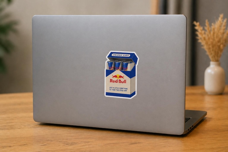 May include: A rectangular sticker on a laptop lid featuring a graphic of a blue and white cigarette pack labelled FRIDGE CIGS. The pack contains small blue and silver energy drink cans and displays the Red Bull logo with the text JUST A LITTLE SOMETHING TO TAKE THE EDGE OFF.