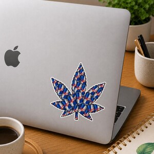 May include: A white sticker in the shape of a cannabis leaf, composed of numerous Red Bull energy drink cans. The sticker is affixed to the lid of a silver laptop, with a coffee cup and notebook in the background.