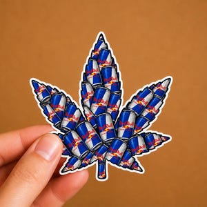 May include: A sticker in the shape of a cannabis leaf, composed of numerous Red Bull energy drink cans. The cans are blue and silver with the Red Bull logo. The sticker has a white border and is held against a brown background.
