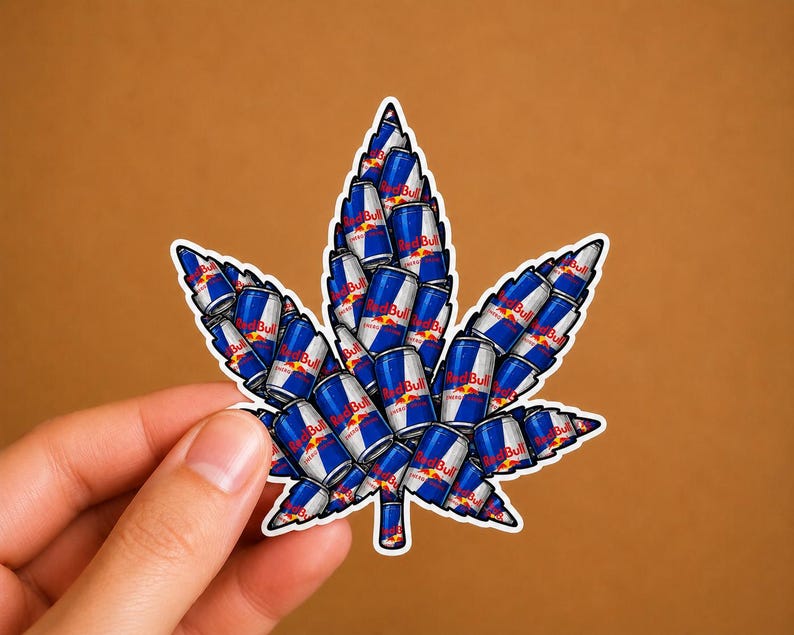 May include: A sticker in the shape of a cannabis leaf, composed of numerous Red Bull energy drink cans. The cans are blue and silver with the Red Bull logo. The sticker has a white border and is held against a brown background.