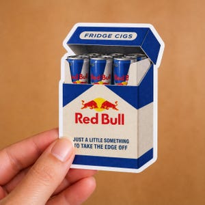 May include: A die-cut sticker shaped like a cigarette pack, featuring a blue and cream colour scheme. The pack is labelled FRIDGE CIGS and Red Bull, with the text JUST A LITTLE SOMETHING TO TAKE THE EDGE OFF. Inside are several blue Red Bull cans.