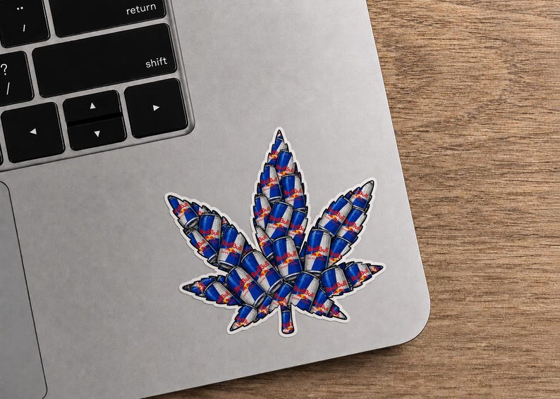 May include: A sticker in the shape of a cannabis leaf, composed of stacked Red Bull energy drink cans. The sticker is on a laptop, with a wooden surface in the background. The cans are blue, silver, red, and white.