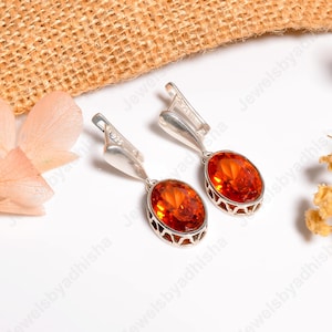 Peut inclure: Une paire de boucles d'oreilles en argent avec des pierres précieuses ovales et orange. Les boucles d'oreilles ont une fermeture à levier et un cadre décoratif en argent. Le fond comprend du tissu tissé et des accents floraux.