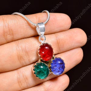May include: A silver pendant necklace featuring three oval-shaped cabochon gemstones arranged in a cluster. The top stone is red, the bottom left stone is green, and the bottom right stone is blue. Each gemstone is set in a scalloped silver bezel.