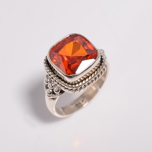 May include: A sterling silver ring featuring a large, square-cut orange gemstone. The ring has a detailed, braided design around the gemstone and a smooth band. The ring is a piece of jewelry.