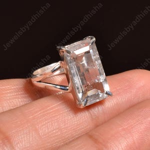 Pode incluir: Anel com uma pedra preciosa transparente de corte retangular e uma banda prateada. A pedra é grande e transparente, engastada num design simples e elegante. O anel é mostrado sobre um fundo claro.