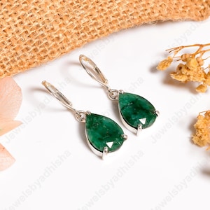 Puede incluir: Un par de pendientes colgantes de plata con piedras preciosas verdes vibrantes en forma de pera. Los pendientes tienen un cierre de palanca y están colocados sobre un fondo blanco con detalles florales.