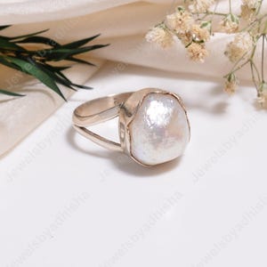Puede incluir: Un anillo de plata con una perla barroca blanca grande e irregular engastada en un bisel de metal liso. La banda del anillo está dividida en dos segmentos de plata distintos que se unen en la base, creando un aspecto abierto y superpuesto.