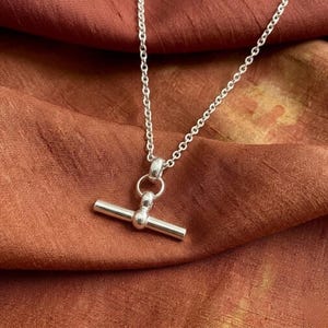 May include: A silver-toned necklace with a T-bar pendant. The necklace chain is delicate, and the pendant features a horizontal bar connected to a vertical bar with a small ball in the center. The necklace is displayed on a textured, rust-colored fabric.