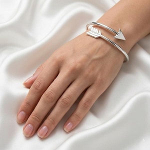 May include: A silver arrow cuff bracelet. The bracelet wraps around the wrist with an arrow design on each end. The bracelet is displayed on a person's wrist against a white satin background.