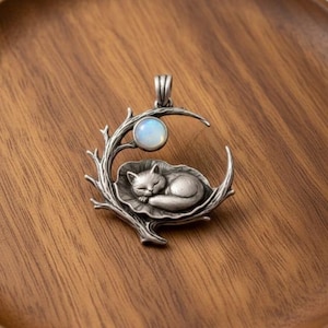 Puede incluir: Colgante plateado con un gato durmiendo en una hoja, enmarcado por una luna creciente y ramas. Una piedra opalina se encuentra sobre el gato. El colgante se muestra sobre una superficie de madera.