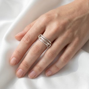 May include: A silver ring with a distinctive design, featuring three interlinked bands. The ring is shown on a hand, highlighting its polished finish and contemporary style. This piece of jewellery is a fashionable accessory.