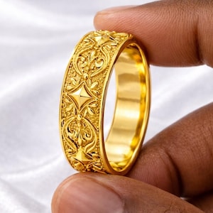 Puede incluir: Un anillo de oro con un diseño ornamentado y texturizado. El anillo presenta un patrón repetitivo de formas florales y geométricas. El anillo se sostiene entre dos dedos, mostrando sus intrincados detalles y su acabado brillante.