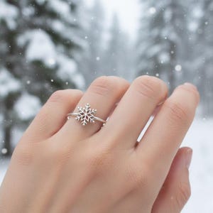 May include: A silver snowflake ring. The ring features a detailed snowflake design, with delicate lines and cutouts. The ring is worn on a finger, set against a snowy, blurred background of trees and falling snow.