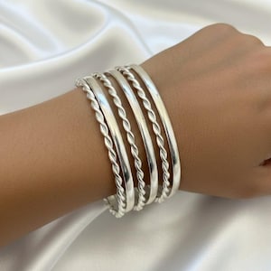 May include: A set of six silver bangles on a wrist. The bangles alternate between a smooth, flat design and a twisted rope design. The jewelry is displayed against a white, silky background.
