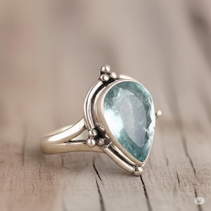May include: A silver ring featuring a large, pear-shaped, light blue gemstone. The ring has a detailed, ornate setting with small decorative beads. The band is split at the base, adding to the ring's unique design.