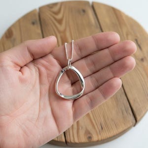 Könnte beinhalten: Eine silberne Halskette mit einem Anhänger in abstrakter Tropfenform. Der Anhänger hängt an einer feinen Silberkette. Die Halskette wird vor einem hellen Holzgrund gezeigt.
