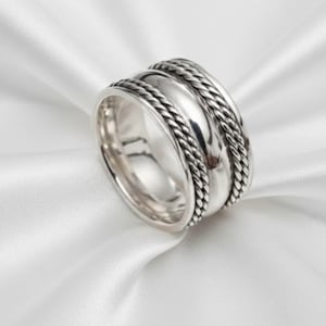 May include: A wide silver ring with a braided rope design on the edges and a smooth, polished centre band. The ring is set against a white, silky background, highlighting its reflective surface.