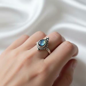 May include: A silver ring featuring a round, blue-green gemstone. The ring has an intricate design around the stone and on the band. The gemstone displays an iridescent shimmer. The ring is worn on a finger.