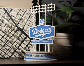 Könnte beinhalten: Ein blau-weißes Dodgers-Baseballstadion-Schild mit dem Logo und Namen des Teams. Das Schild steht auf einer weißen Basis mit den Worten "LOS ANGELES BASEBALL" und "STADIUM - LOS ANGELES, CA" aufgedruckt. Im Hintergrund befindet sich eine weiße Karte der Stadt.