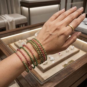 Op de afbeelding: Een stapel van vijf dunne gouden armbanden met afwisselend kleine ronde groene en roze edelstenen, gedragen naast een eenvoudige gouden ring. De armbanden zijn in een afwisselende kleurvolgorde om de pols gerangschikt.