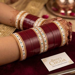 Puede incluir: Un conjunto de pulseras de novia granates y blancas. Las pulseras están apiladas en la muñeca, con una banda blanca con detalles de puntos de colores y cuentas doradas. Se ve una tarjeta de visita con el texto "Bride's Chuda".