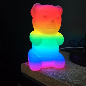 May include: A rainbow-colored teddy bear-shaped lamp. The lamp is illuminated with a gradient of colors, transitioning from red at the top to blue at the bottom. The lamp is sitting on a wooden surface.