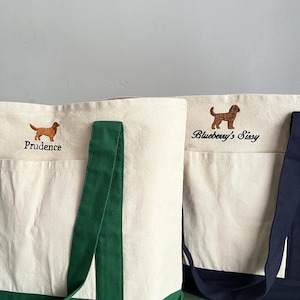 K&ouml;nnte beinhalten: Zwei Canvas-Tragetaschen mit gestickten Hundeillustrationen. Eine Tasche hat einen gr&uuml;nen Boden und Henkel, die andere einen marineblauen Boden und Henkel. Auf der ersten Tasche ist der Name "Prudence" &uuml;ber dem Hund gestickt. Auf der zweiten Tasche steht "Blueberry's Sissy".