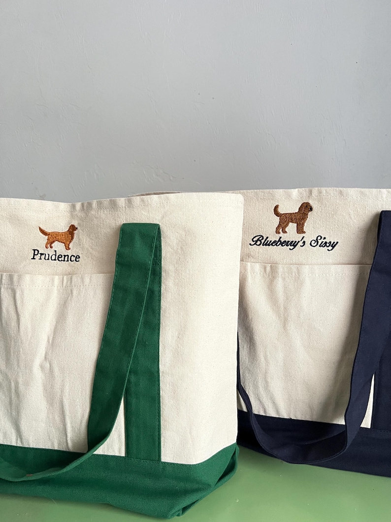 K&ouml;nnte beinhalten: Zwei Canvas-Tragetaschen mit gestickten Hundeillustrationen. Eine Tasche hat einen gr&uuml;nen Boden und Henkel, die andere einen marineblauen Boden und Henkel. Auf der ersten Tasche ist der Name "Prudence" &uuml;ber dem Hund gestickt. Auf der zweiten Tasche steht "Blueberry's Sissy".