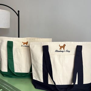 K&ouml;nnte beinhalten: Zwei Canvas-Tragetaschen mit gestickten Hunde-Designs. Eine Tasche hat gr&uuml;ne Griffe und eine Tasche, mit dem Namen "Prudence" &uuml;ber der Tasche gestickt. Die andere Tasche hat marineblaue Griffe und den Namen "Blueberry's Sissy" darauf gestickt.