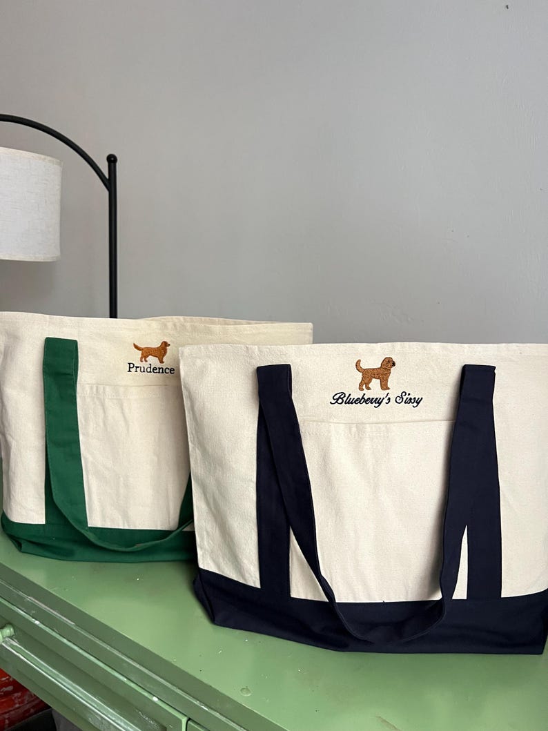 K&ouml;nnte beinhalten: Zwei Canvas-Tragetaschen mit gestickten Hunde-Designs. Eine Tasche hat gr&uuml;ne Griffe und eine Tasche, mit dem Namen "Prudence" &uuml;ber der Tasche gestickt. Die andere Tasche hat marineblaue Griffe und den Namen "Blueberry's Sissy" darauf gestickt.