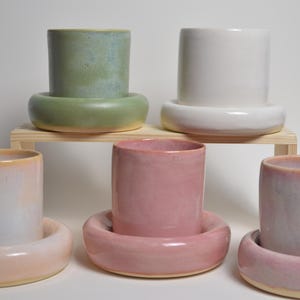May include: A selection of ceramic cups in shades of green, white, pink, and peach. Each cup rests on a matching, rounded base. Some are displayed on a small wooden shelf. The cups have a smooth, glossy finish.