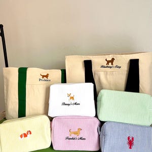 K&ouml;nnte beinhalten: Eine Auswahl an Canvas-Tragetaschen und Kosmetiktaschen mit gestickten Hunde- und Hummer-Designs. Die Taschen weisen verschiedene Farben und Streifenmuster auf, mit Namen wie "Prudence" und "Frankie's Mom" aufgestickt.