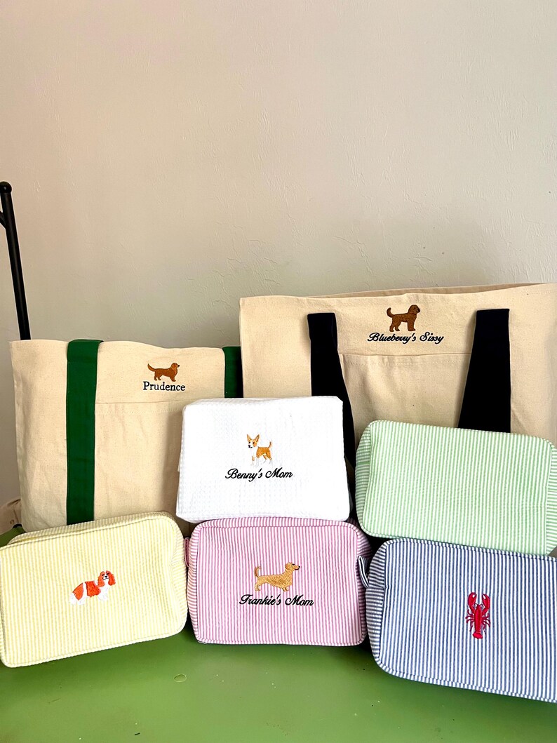 K&ouml;nnte beinhalten: Eine Auswahl an Canvas-Tragetaschen und Kosmetiktaschen mit gestickten Hunde- und Hummer-Designs. Die Taschen weisen verschiedene Farben und Streifenmuster auf, mit Namen wie "Prudence" und "Frankie's Mom" aufgestickt.