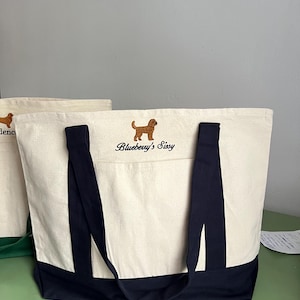 K&ouml;nnte beinhalten: Cremefarbene Tragetasche mit marineblauen Akzenten und Griffen. Die Tasche hat einen gestickten braunen Hund und den Text "Blueberry's Sissy". Aus robustem Canvas-Material.