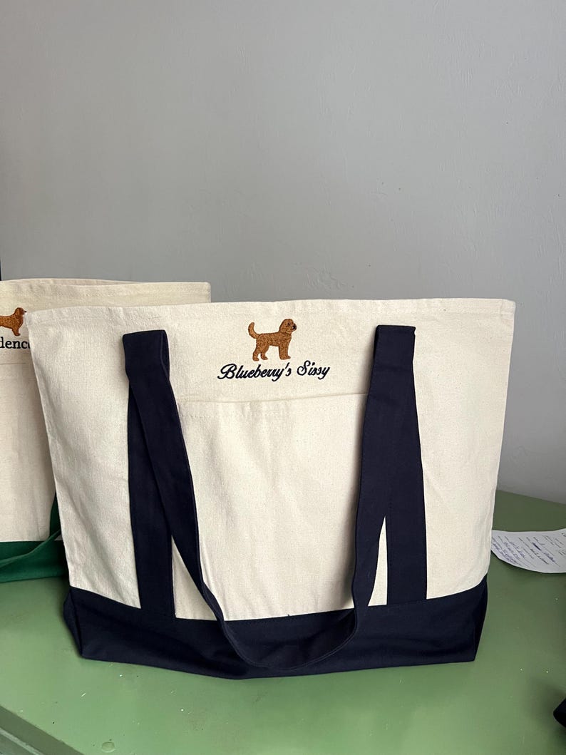 K&ouml;nnte beinhalten: Cremefarbene Tragetasche mit marineblauen Akzenten und Griffen. Die Tasche hat einen gestickten braunen Hund und den Text "Blueberry's Sissy". Aus robustem Canvas-Material.