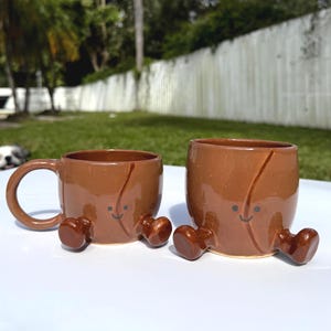 May include: Two brown ceramic mugs with a whimsical design. One mug has a handle, and both have small, rounded feet and a smiling face. The mugs are set on a white surface against a blurred outdoor background.