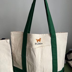 K&ouml;nnte beinhalten: Cremefarbene Canvas-Tragetasche mit dunkelgr&uuml;nen Akzenten. Mit einer Vordertasche und dunkelgr&uuml;nen Griffen. Ein gestickter Golden Retriever Hund und der Name "Prudence" sind auf der Vorderseite. Die Tasche wird von einer Person gehalten.