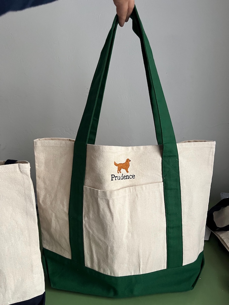 K&ouml;nnte beinhalten: Cremefarbene Canvas-Tragetasche mit dunkelgr&uuml;nen Akzenten. Mit einer Vordertasche und dunkelgr&uuml;nen Griffen. Ein gestickter Golden Retriever Hund und der Name "Prudence" sind auf der Vorderseite. Die Tasche wird von einer Person gehalten.