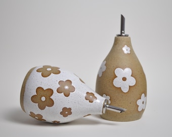 May include: Two ceramic oil and vinegar dispensers. One is lying on its side, the other upright. Both have a teardrop shape, a stainless steel spout, and a floral pattern. The upright dispenser is tan with white flowers.
